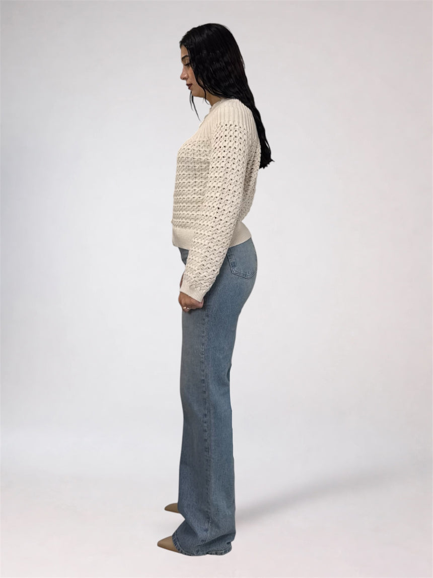 A woman shown in profile wearing a cream open-knit cropped sweater and light blue flared jeans against a plain light gray studio background.