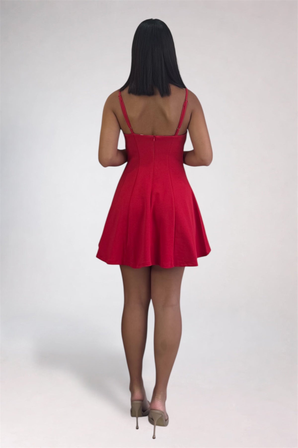 A woman viewed from behind wearing a bright red sleeveless mini dress with thin straps and a flared skirt, paired with nude high heels, against a plain light gray studio background.