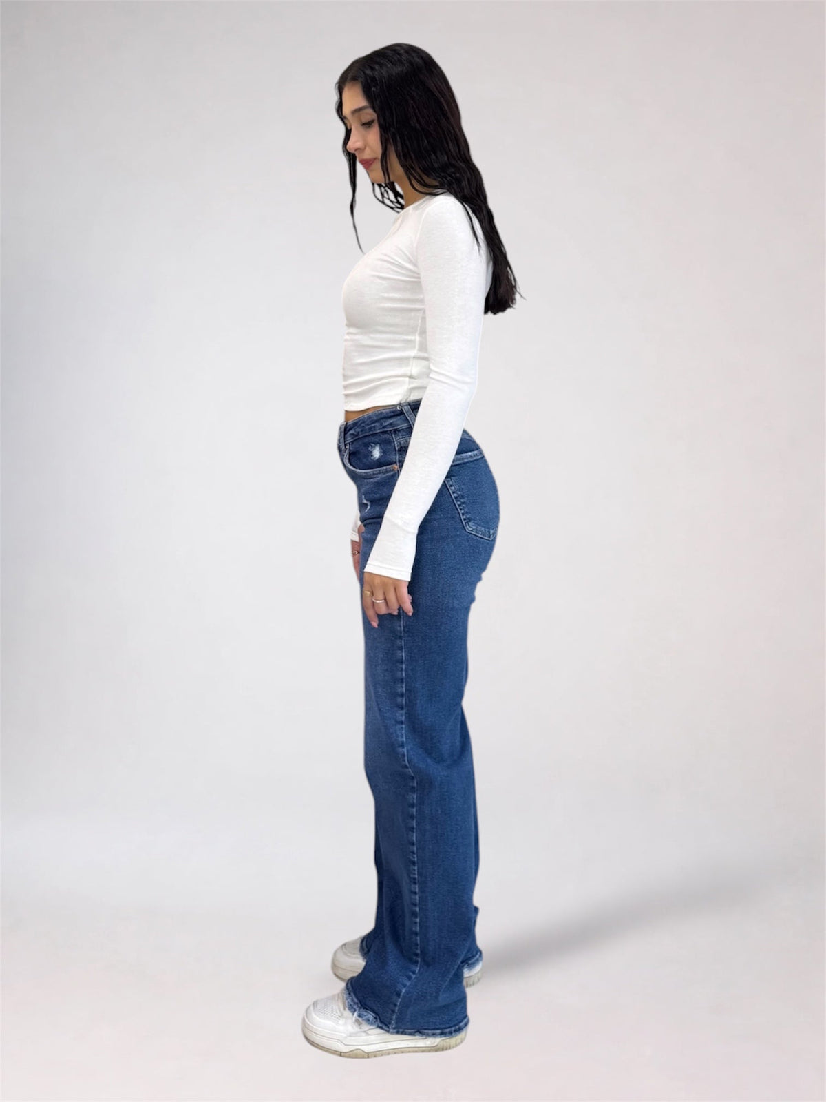 A woman standing in profile wearing a fitted white long-sleeve top, blue high-waisted flare jeans, and white sneakers against a plain light gray studio background.