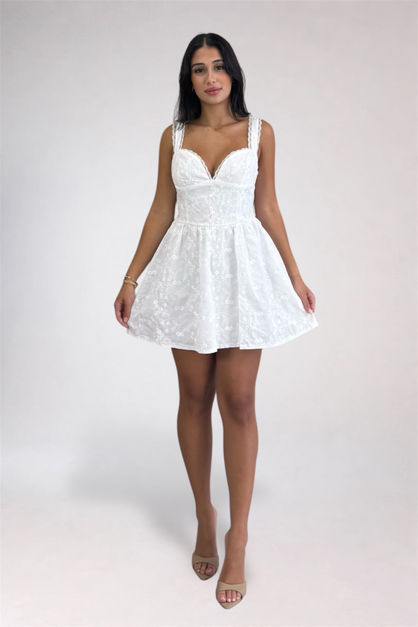 A woman modeling a short white lace dress with a sweetheart neckline, thin shoulder straps, and a flared skirt, photographed against a plain light gray studio background.