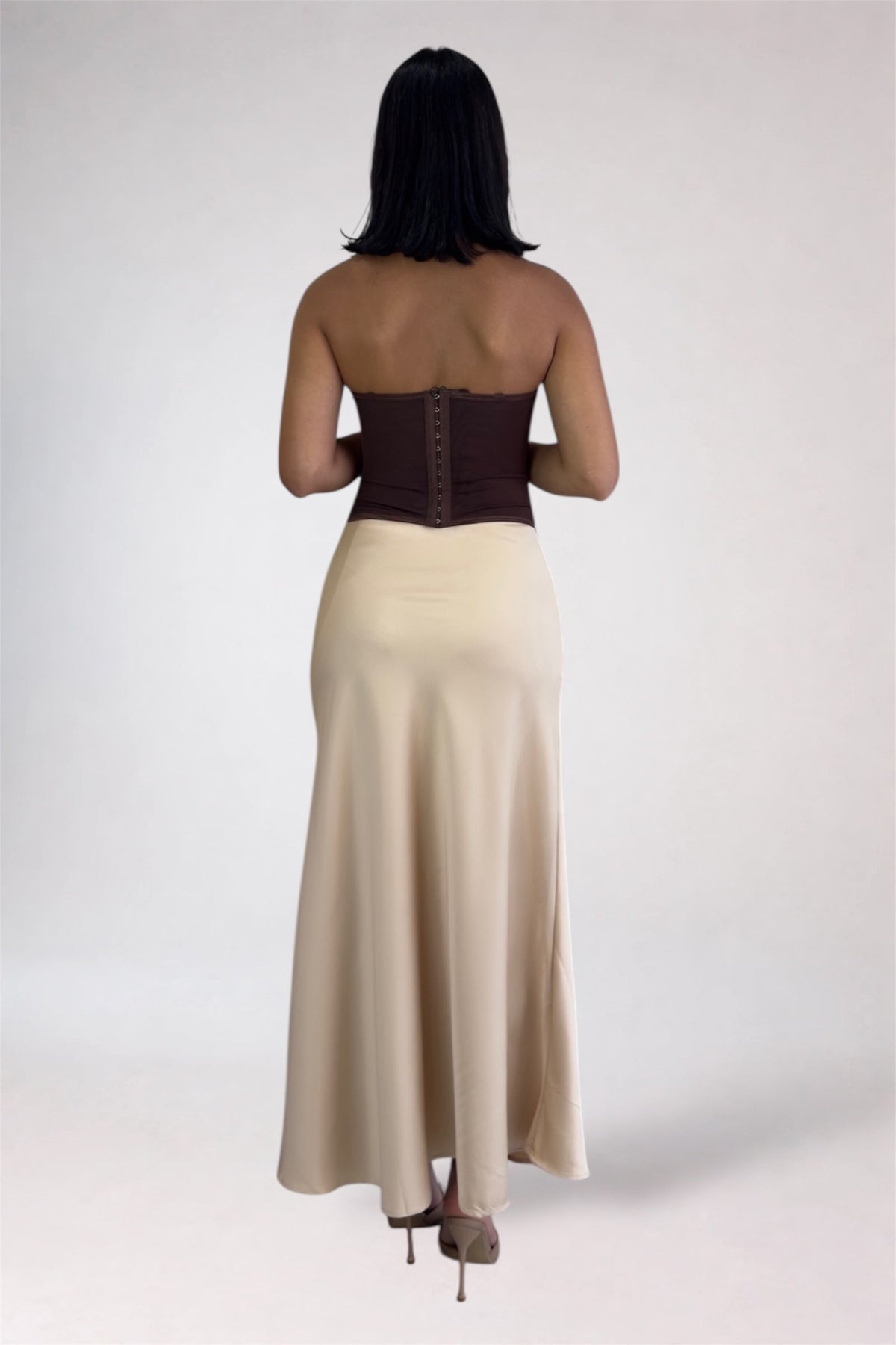 A woman shown from the back wearing a strapless maxi dress with a dark brown corset-style upper back and a flowing cream skirt, photographed against a light gray studio background.