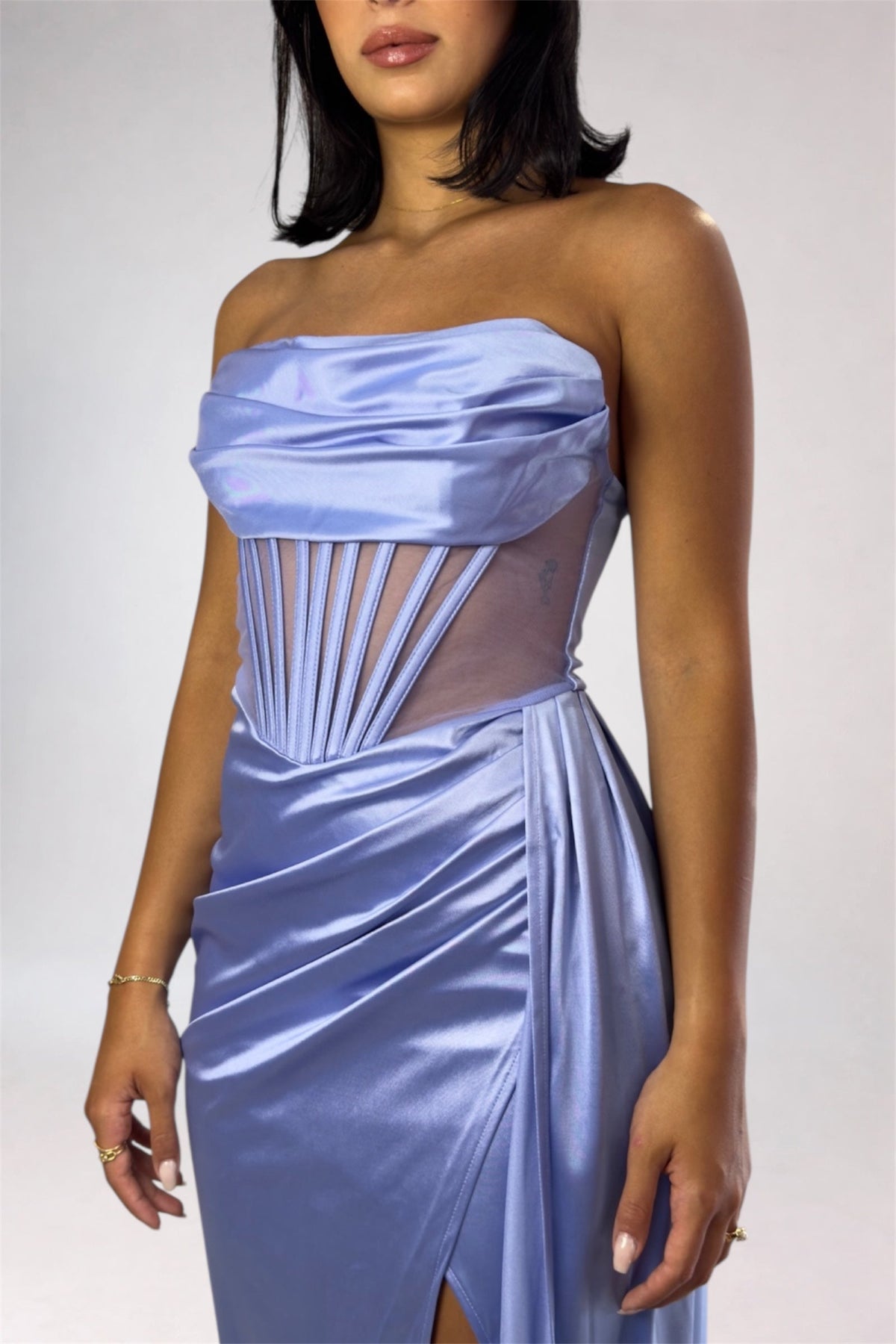 A woman wearing a strapless light blue satin gown with a ruched bodice, sheer side panels, and a draped skirt, shown against a plain light gray studio background.