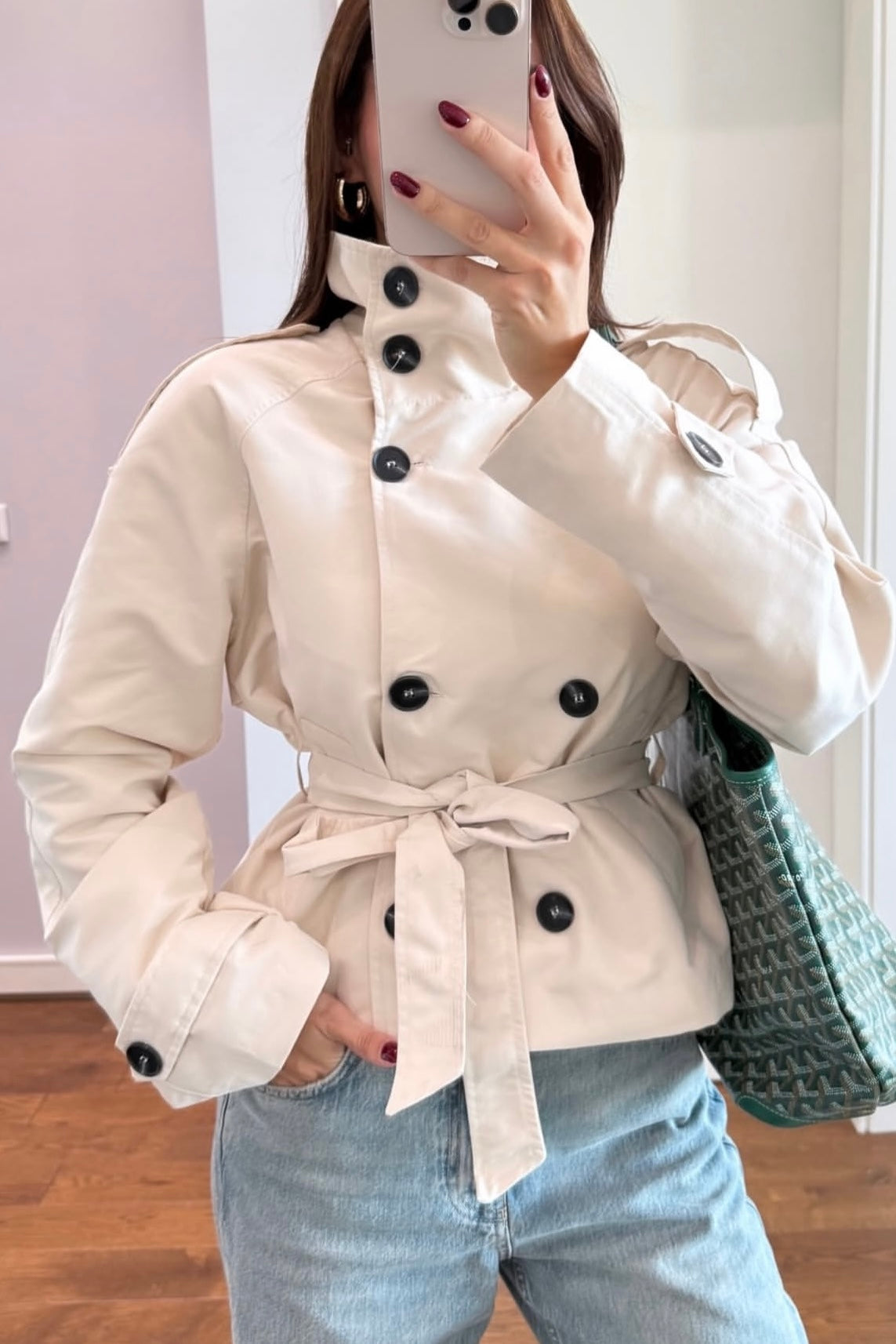 Person wearing a beige trench coat with black buttons, holding a phone and a green bag indoors.