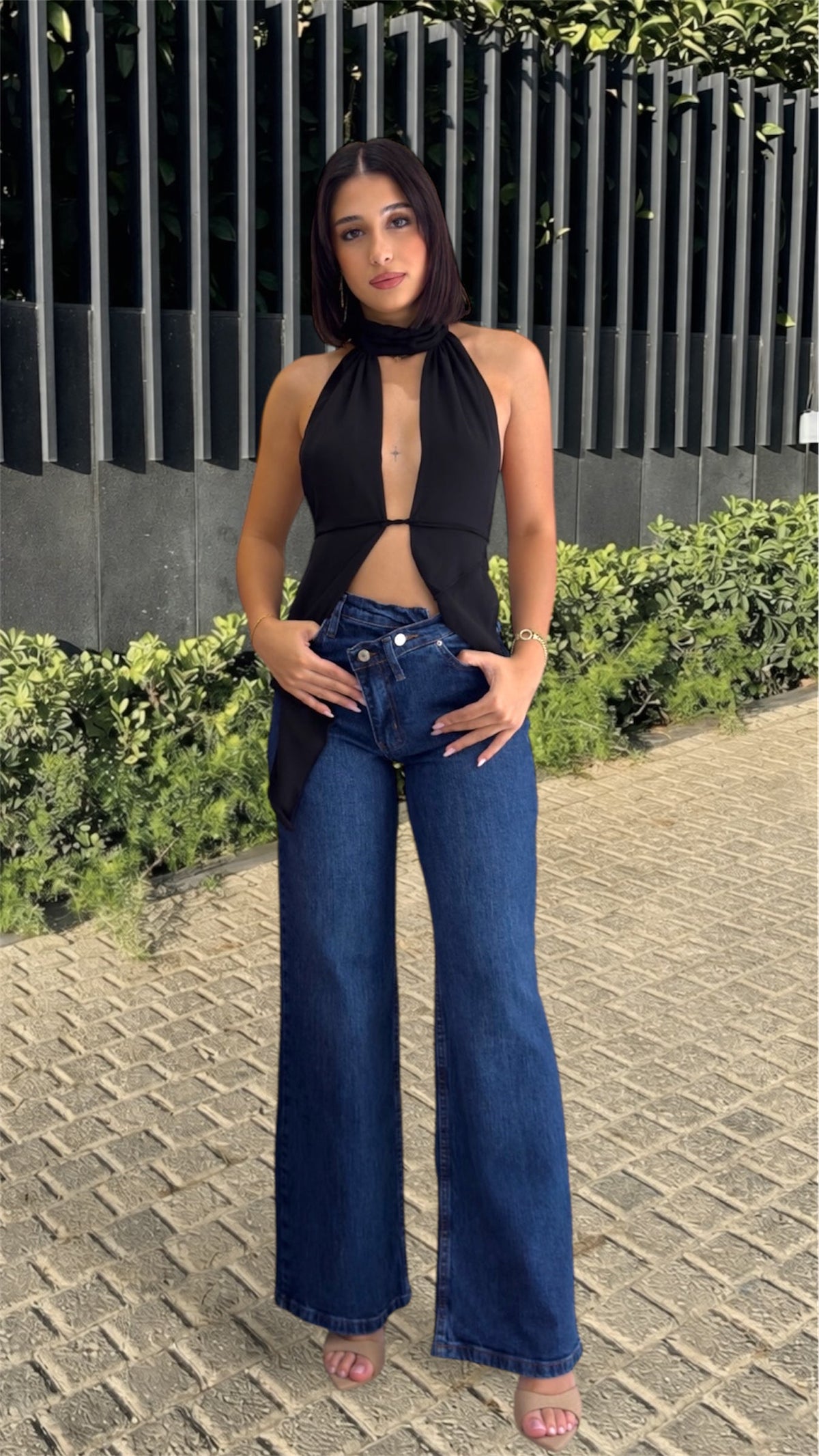 Woman wearing a black halter top and blue jeans standing outdoors on a paved area with greenery.