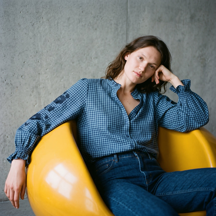 Woman wearing an KIDDING indigo blue gingham check blouse with floral embroidered sleeves and blue jeans.