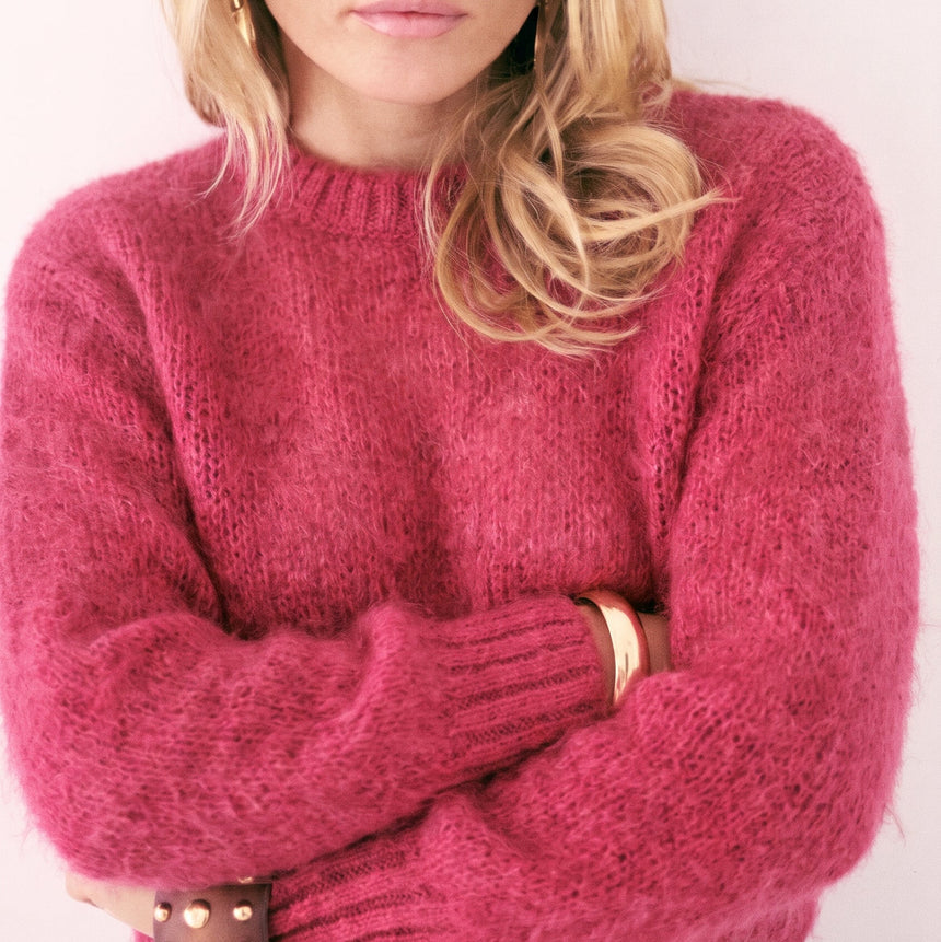Close-up of a blonde woman wearing a KIDDING vibrant raspberry pink alpaca-blend crewneck JUMPER with ribbed cuffs and statement black sunglasses.