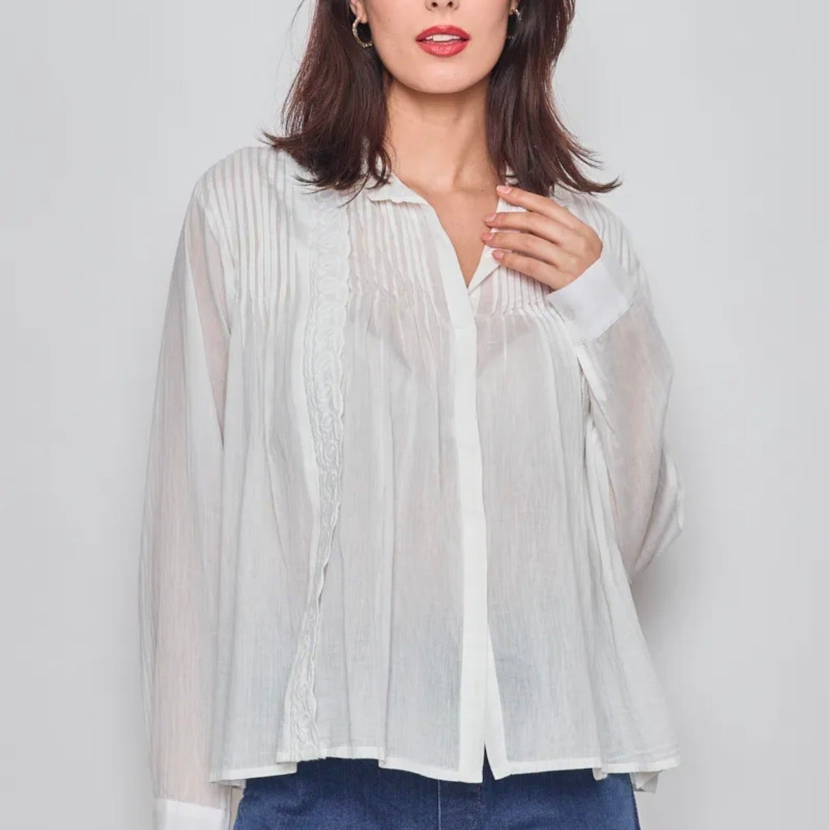 Woman wearing a KIDDING white semi-sheer long-sleeve blouse with pintuck pleats and lace trim details paired with blue jeans, posing against a light grey background