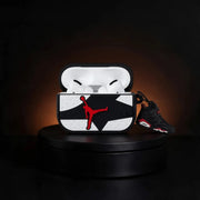 A custom AirPods case featuring a black and white geometric pattern with a red Jordan logo, shown open with white AirPods inside and a black and red Air Jordan sneaker resting beside it on a black display pedestal.