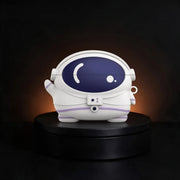 A white astronaut-shaped case for airpods and buds,  with a dark blue visor and subtle purple accents, displayed on a black circular pedestal against a dark background with warm light glow.