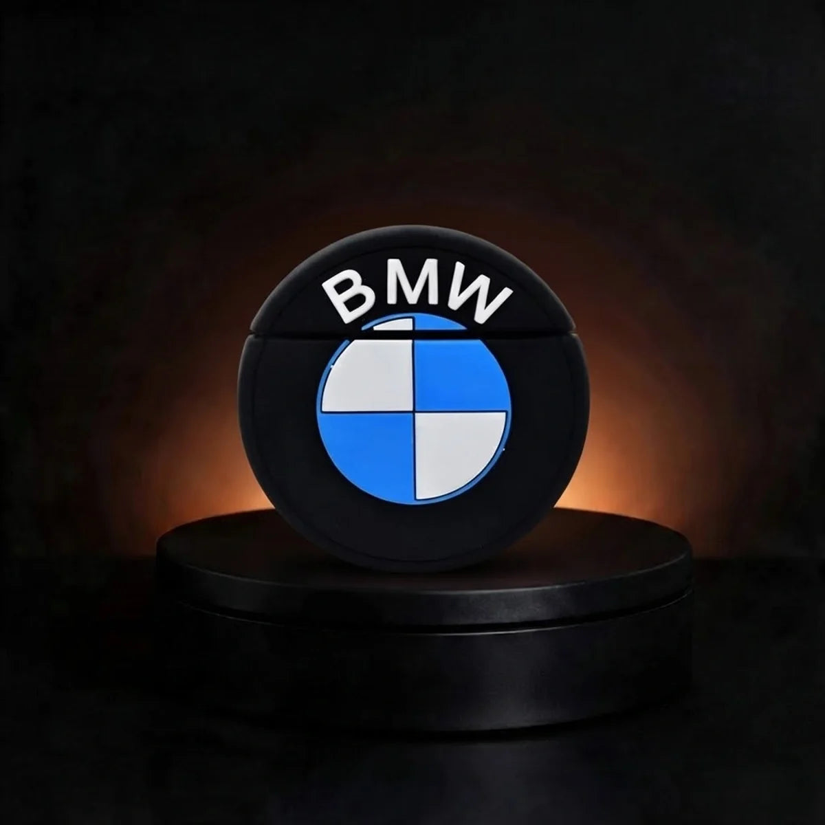 A circular black BMW logo case for airpods featuring the white BMW wordmark and blue-and-white quadrilobe emblem, displayed on a black pedestal against a dark background with warm light glow.