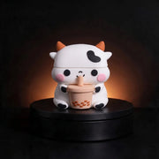 A cute cartoon cow airpods case with orange horns and a black heart-shaped patch on its forehead, holding a cup of bubble tea. The figurine is displayed on a black circular pedestal against a dark background with a warm glow behind it.