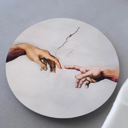 A round coaster with an engraved image of Michelangelo's 'The Creation of Adam' painting on a light background, placed next to a white cloth.