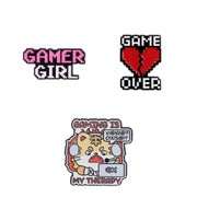 Three gaming-themed enamel pins on a plain white background: a pink and white pixel-style 'GAMER GIRL' pin, a red broken-heart pixel pin with black text reading 'GAME OVER,' and a multicolor cartoon pin showing a tiger character holding a laptop with the text 'GAMING IS MY THERAPY' and a speech bubble.