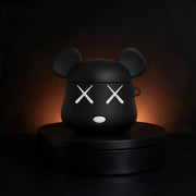 A matte black bear-shaped case for airpods, resembling a stylized character head with mouse ears, white X eyes, and a small white oval mouth, displayed on a round black pedestal against a dark background with warm orange glow behind it.