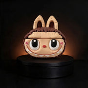 A stylized cartoon character airpods case or decorative object with large eyes, a small red nose, pink cheeks, and brown hair, displayed on a black round pedestal against a dark background with warm light glow.