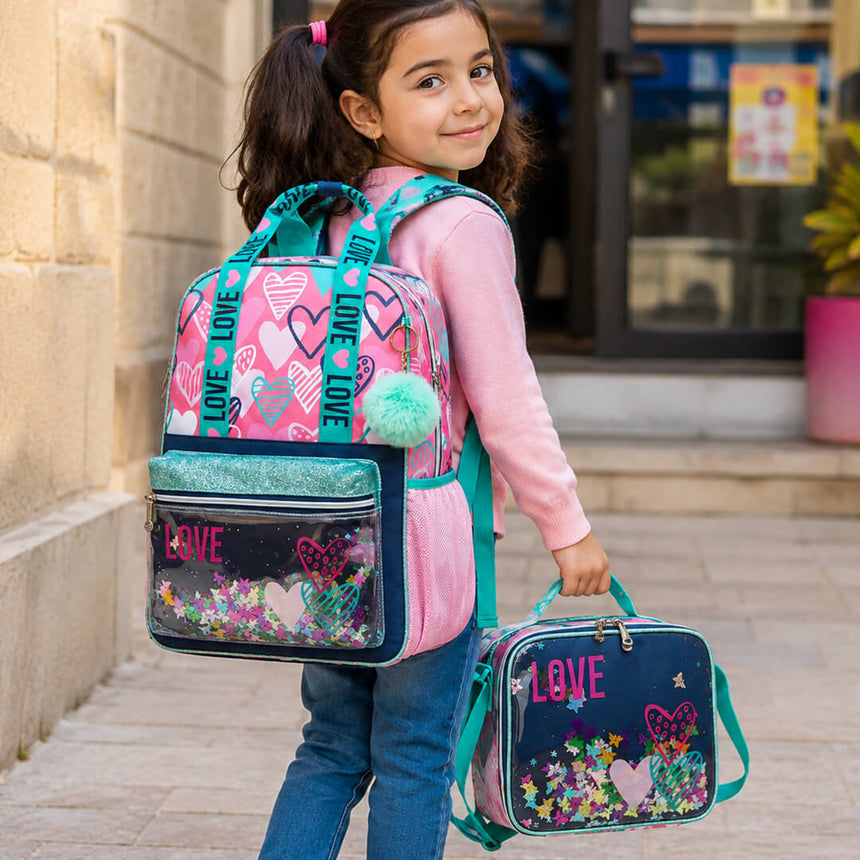 A young girl wearing a pink and teal backpack and holding a matching lunch bag with glittery heart designs