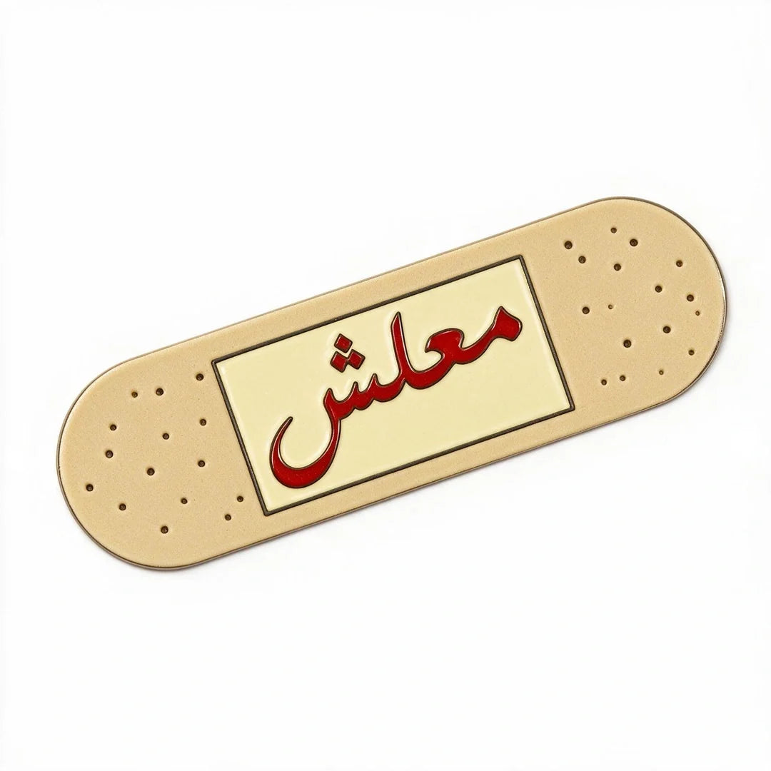 A novelty enamel phone sticker shaped like a beige band-aid with red Arabic calligraphy spelling 'معلش' (Maalesh) on a cream center panel, set against a plain white background.