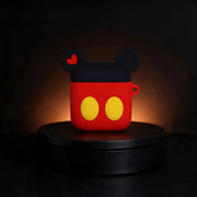 A red and black silicone AirPods case designed like Mickey Mouse's head, featuring yellow circular eyes and a small red heart on the ear, displayed on a black circular pedestal against a dark background.