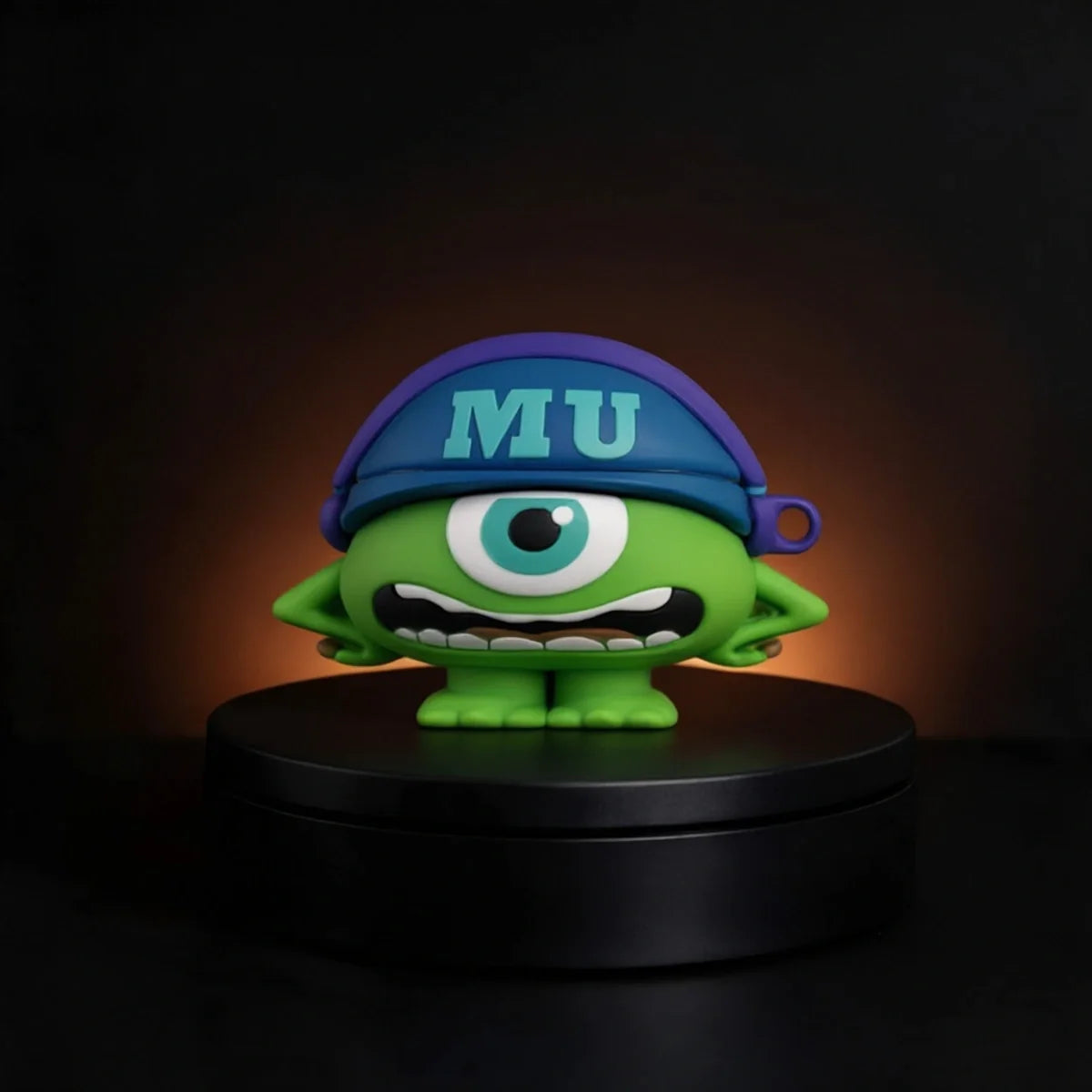 A green one-eyed cartoon character mike wazowski from monsters inc airpods and buds case figurine wearing a blue cap with 'MU' in light blue, standing on a black circular base against a dark background with a warm glow.