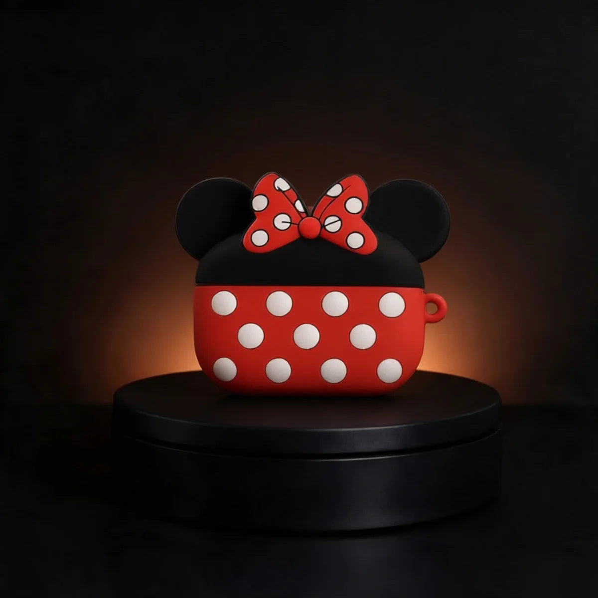 A red and black AirPods case designed like Minnie Mouse's head, featuring white polka dots and a red bow with white polka dots, displayed on a black circular pedestal against a dark background.