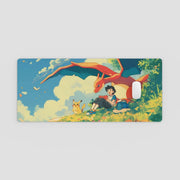Desk Mat with an illustration of a person sitting with two dragons and a bird on a grassy hill with a blue sky.