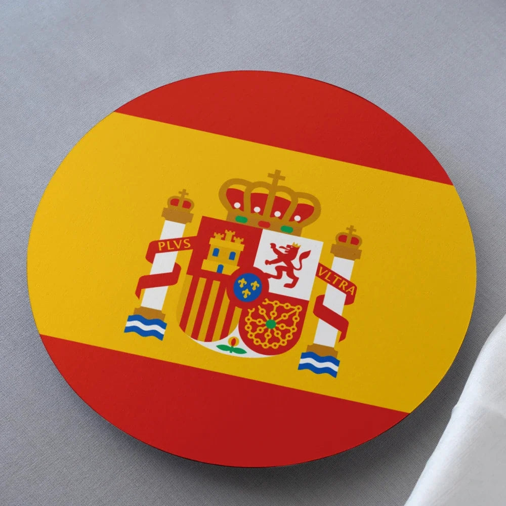 A round coaster featuring the Spanish flag design with its red and yellow horizontal bands and central coat of arms, photographed on a neutral gray surface.