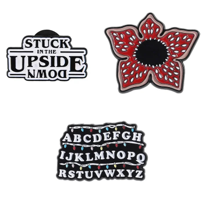 Three decorative pins on a white background: 'Stuck in the Upside Down', a red flower-like design, and a letter pin.