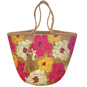 A beige-colored straw tote beach bag with multicolored floral patterns and a tassel detail.